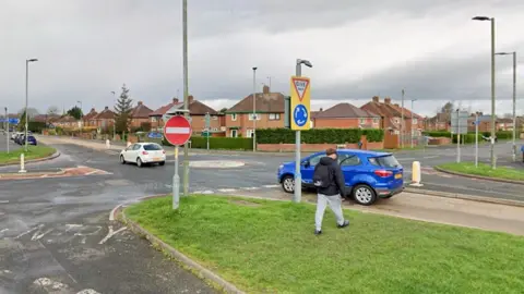 Google The Holme Lacy Road in Hereford. Cars are crossing a mini-roundabout while someone is walking on a grass verge on the side of the road.