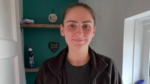 Woman wearing black BCP fleece in a bathroom with bleach on shelf behind smiling at camera
