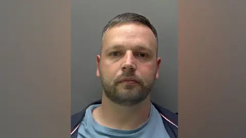 National Crime Agency Irakli Rustemi has short brown hair, a mustache and a beard. He is looking at the camera and is wearing a light blue t-shirt and a dark blue jacket.