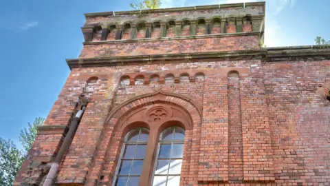 Ray Cartwright A close-up view of an old brick mill building with arched windows and plants sprouting from the roof