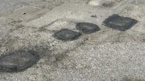 Four potholes filled in with asphalt on a road. They go from bottom left to top right on the image.