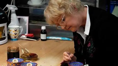 A worker paints the finish on chinaware in a pottery in Stoke-on-Trent. She is leaning forward and using a paintbrush. Paint and china are on the desk in front of her. She is wearing glasses and concentrating.