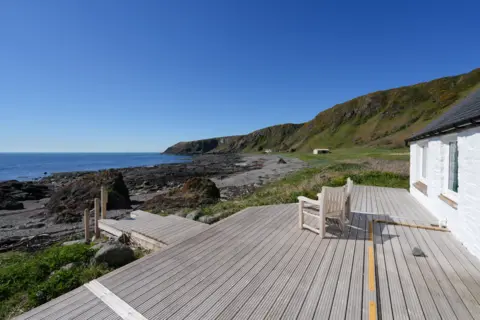 Galbraith Two wooden seats on a large expanse of decking looking out over a bay and towards the sea