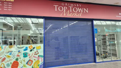 A shop unit with floor to ceiling glass windows either side a blue shuttered entrance. Above is a burgundy sign with 'Grimsby Town Town Market' in white lettering in the middle, on the left are opening times also in white type face.