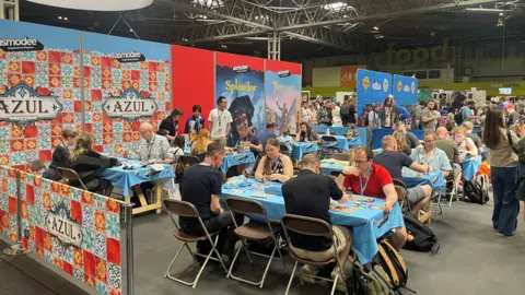 A lot of people sitting at tables playing board games at an exhibition. There are people standing around them and advertising boards have been put up next to them.