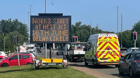 Emma Craig/BBC A black, electronic sign by the side of a busy road. Orange writing says that works start at the next junction on the 16th of June and that delays are likely.