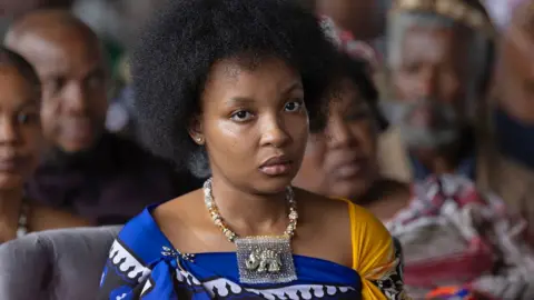 SIHLE MDLULI is provided to the camera in a dangerous expression. She wears her hair in a style of Afro and has a thick necklace of beads with the design of the elephant around her neck and wears yellow and blue outfit.