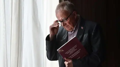 Getty Images Professor Phil Scraton is seen clutching a report, he has white hair and is holding his glasses.