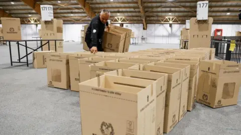An election worker with a dark jacket arranges cardboard ballot boxes in a large hall