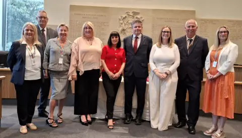 Thurrock Council Nine people, a mix of women and men, stand in a line facing the camera and standing in the Thurrock Council chamber. Lynn Worrall, wearing a red top, is standing in the centre, and John Kent, wearing a suit and red tie, is standing to her left.