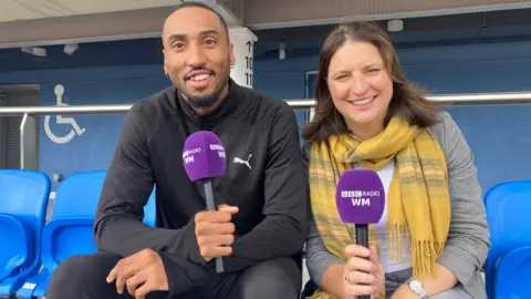 A man wearing a dark tracksuit and a woman wearing a yellow scarf and grey jacket, smile while holding purple BBC Radio WM microphones.