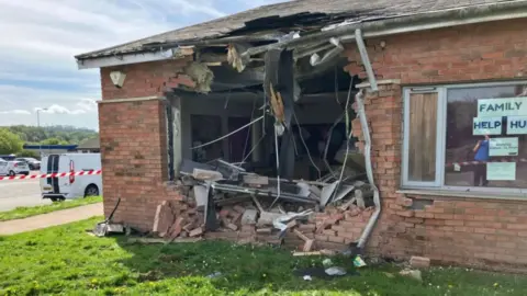 A one-storey brick building with a large hole knocked in one wall and red and white tape to the left