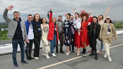 Sandra Zhukova A gathering of people in traditional dress with their arms in the air, standing on a road with trees in the distance behind them.