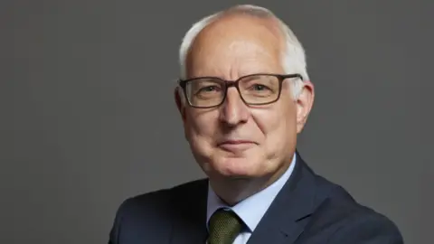 UK Parliament A man with glasses and white hair smiling into the camera. He is wearing a suite and tie.