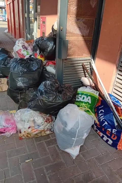 LDRS Piles of black and white rubbish bags and loose waste stacked along a narrow pavement outside a building entrance, partially blocking a doorway and access panel.