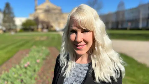 Claire Cox, who has bleached blonde and wavy hair smiles at the camera. She is standing in some gardens, which are blurred out in the background. She is wearing a black jacket with a smart black and white top underneath.