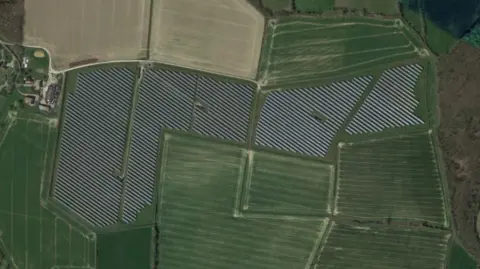 A bird's eye view of a solar farm. The farm includes lots of black solar panels surrounded by green and cream-coloured fields