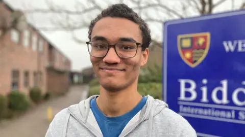 Alex Pope/BBC Stan, standing outside a school, by a school sign. He has short dark hair, is wearing glasses and has on a grey hoodie and blue T-shirt. He is looking straight at the camera. Trees and buildings are behind him.