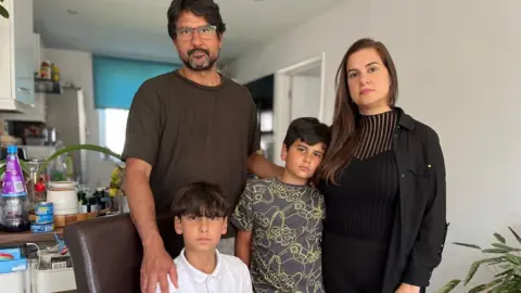 Hugo in a dark olive T-shirt and Ana in a black top with their sons Luca (centre) in a grey and yellow patterned T-shirt and Guilherme (seated) in a white polo shirt with a kitchen in the background. All four of them have serious expressions.