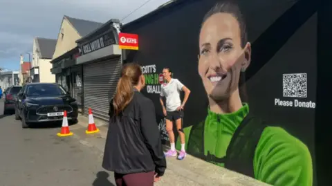 Jill Scott stands in front of a mural of her face on a wall in a street in Sunderland. Jill wears a white shirt and black shorts and her mural has her wearing a green fleece and black backpack against a black background. 