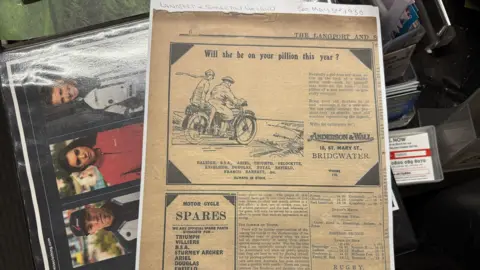 A newspaper clipping from 1930 on top of a photo album. The newspaper has an advert for Anderson & Wall in it.