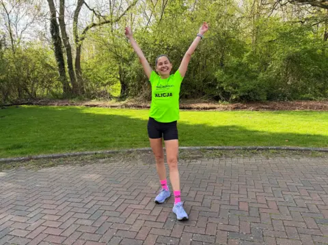 Alicja Bunio A woman is stood with her arms raised above her head, smiling widely at the camera. She is in a bright green top, black shorts, bright pink socks and pale purple trainers.