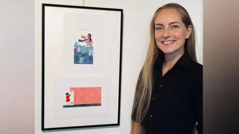 Paula Heister has long blonde hair and is standing next to some of her artwork. She is wearing a black shirt and is smiling.