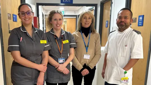 Royal United Hospitals Bath Four people in scrubs are standing in a row, some with their arms crossed in front of them. They are standing in a hospital hallway. They are all smiling at the camera.