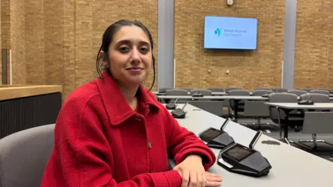 Martin Heath/BBC Shanika Mahendran, sitting at a desk, wearing a red jacket, in a council room, with an audio machine in front of her. The walls are brick, and there is a television screen on the wall. There are chairs and tables in the room. 