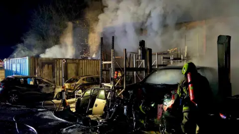 South Yorkshire Fire & Rescue A firefighter stands in front of a smouldering collection of cars and metal racks. It is dark but smoke car be seen coming from the debris. There are large pools of water on the floor.