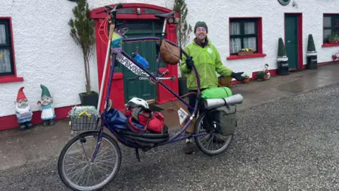 A man is standing beside a large bicycle. The seat and handlebars are taller than his head. The bicycle is painted purple and has baskets and bags attached on the front and back. The man is wearing a green coat and hat. 