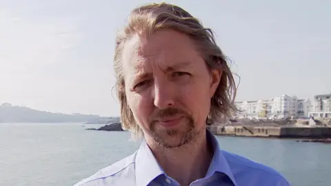 Councillor Josh McCarty stood on the waterfront at Plymouth Hoe on a bright sunny day. The sea is in the background and is calm with some buildings in the distance. He is look at the camera smiling while wearing a light blue shirt. 