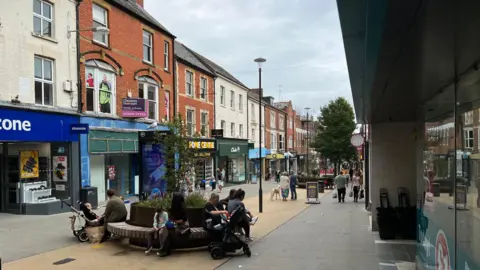 An image of a pedestrianised shopping street, people sit on benches around a planter. Shops are visible in the background. 