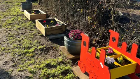 Jyan Dutton Planters containing yellow, white and purple flowers. The one at the front is shaped like a train engine and is orange and yellow in colour. 