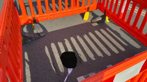 BBC A sinkhole within a square-shaped cordoned-off area surrounded by red barriers.