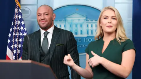 Conor Mc Gregor is standing in front of a US flag and a White House logo. He is wearing a three-piece, pinstripe suit and a dark green tie. His hair and beard are closely cropped and he is smiling. Beside him and gesturing towards him is Karoline Leavitt, she has blonde, shoulder length hair and is wearing a dark green dress.