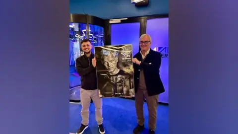 Wilf Elliott Artist Wilf Elliott and Avicii's father Klas Bergling holding up a black and white drawing of the late DJ. The artist is wearing a black zip-up jacket and cream trousers, and Mr Bergling is in a black jacket, white shirt, and beige jumper and trousers. They are in a room that has blue carpet and blue walls.