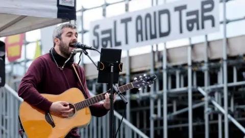 Claire Hamilton/BBC A musician wearing a purple sweater with a black turtle neck shirt plays an acoustic guitar outside Salt And Tar.