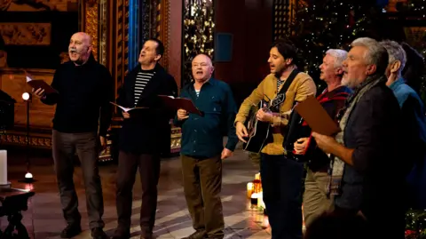 A picture of seven people standing in a line. They are holding books while singing. They are standing in front of decorations and on a wooden floor at Westminster Abbey.
