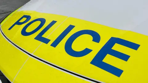 The front of a police vehicle with the word 'Police' on the bonnet in blue. The car is yellow and white.