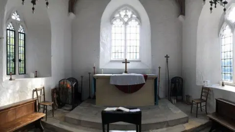 Bill Nicholls The inside of Bartlemas Chapel. There is an altar with a cross on it and a large glass window above it. The chapel interior is rustic, with the large walls broken up by large windows.
