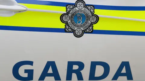Close up of Garda emblem on patrol vehicle door. Shows logo on a bright yellow background, with blue lettering below reading 'GARDA'.