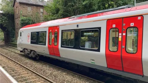 A Transport for Wales train in red and grey livery, on a rail track near a boarded up building