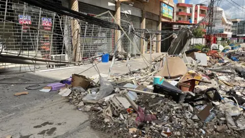 Piles of rubble, cardboard and other items are visible on the ground as a power line has been pulled down. Shops are closed and gated along the street. 