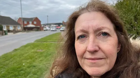 John Devine/BBC Helen Arnold, 56, has shoulder length wavy brown hair swept over her forehead behind her is a busy road with some housing visible and cars parked and in motion all the roadway.