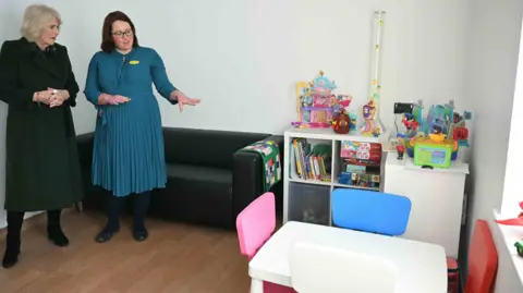 PA Media Queen Camilla (left) is shown a paediatric non-acute room with toys, a table and chairs and other furniture including a bookcase for children.