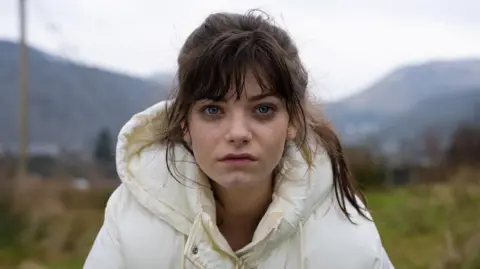 A young woman with brown hair in a ponytail and wearing a white puffer coat stares at the camera with a sad expression