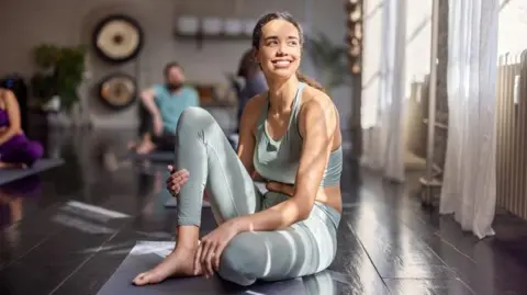 Getty Images A woman with long brown hair tied up is sitting in her pale green exercise leggings and crop top on a grey yoga mat looking happy after a class. Other people are sitting in background. The sunlit room has long white curtains at the windows and wooden floorboards.