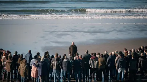 Sky A crowd are gathered on the beach, looking out to sea as Sir Jonathan Pryce's character looks back at them, with the waves behind him.