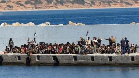 A large crowd of migrants waits on a concrete jetty